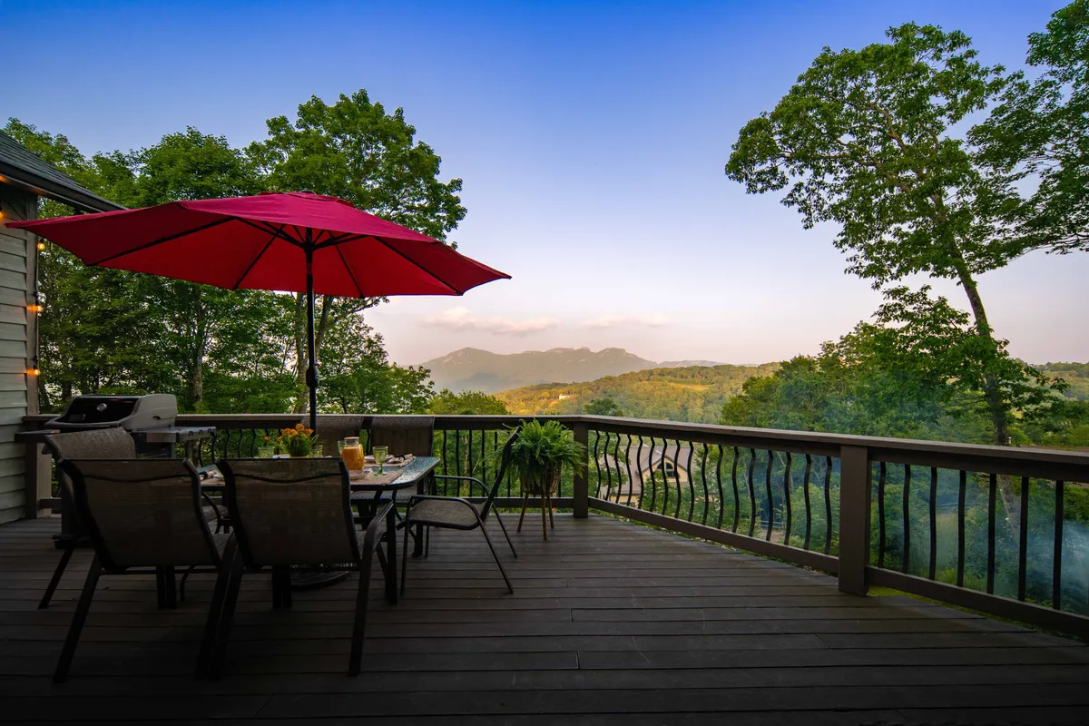 This exterior image captures an expansive deck featuring a red umbrella shading a dining table, surrounded by lush green trees and set against a stunning mountain view. The scene suggests an inviting space for outdoor dining, barbecuing, or simply relaxing and soaking in nature.