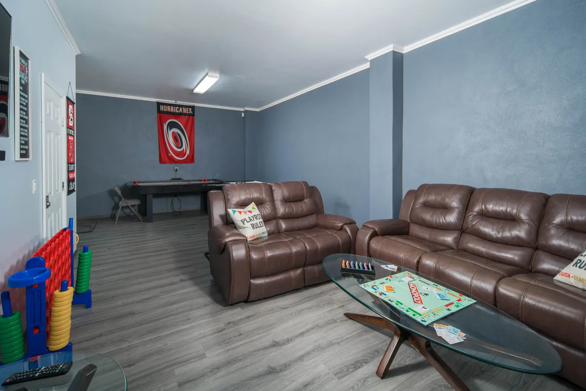 A vibrant playroom featuring brown leather sofas, a glass coffee table with colorful games and toys, an air hockey table, and a team banner on the wall. The setup invites group play and family gatherings, with plenty of space for both relaxation and friendly competition.