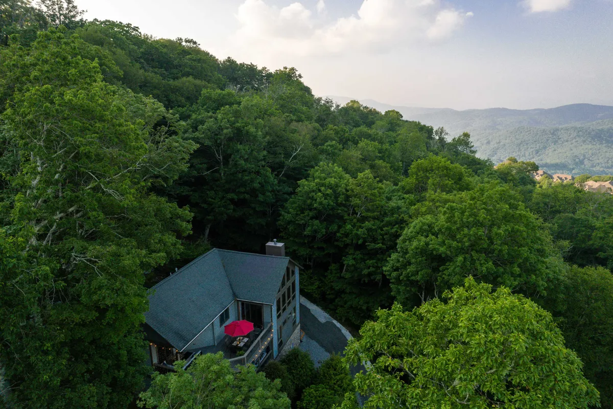 A wide shot of the house nestled in lush greenery with a red umbrella on the deck and a beautiful mountain backdrop. The scene evokes a tranquil, nature-filled atmosphere and showcases the inviting outdoor deck that is perfect for relaxing with a pet after a day of adventure.
