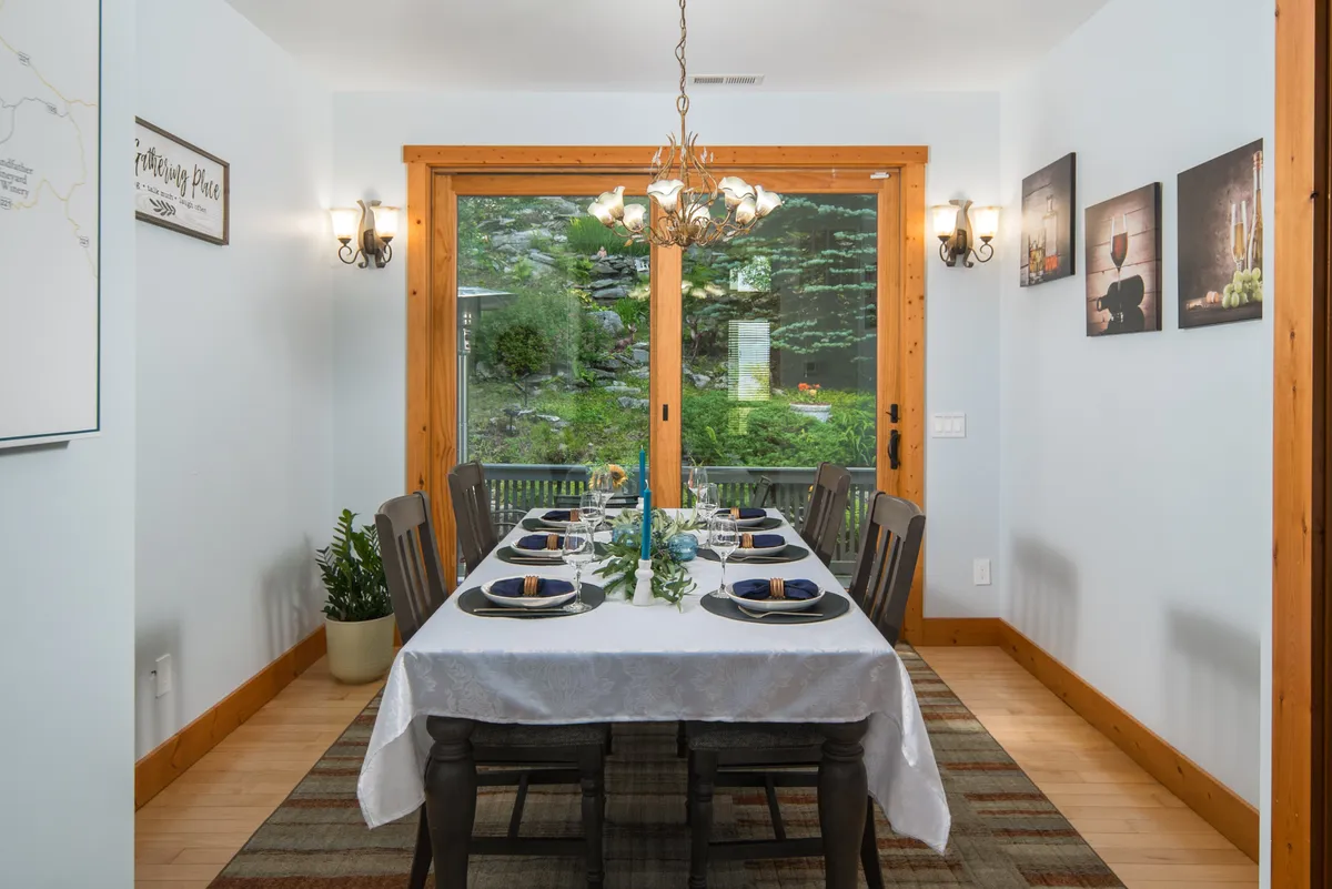 A spacious dining area with a large table set beneath a chandelier, artwork on the walls, and windows letting in natural light and views. The table is perfectly suited for group meals and gathering for celebrations or casual dining.