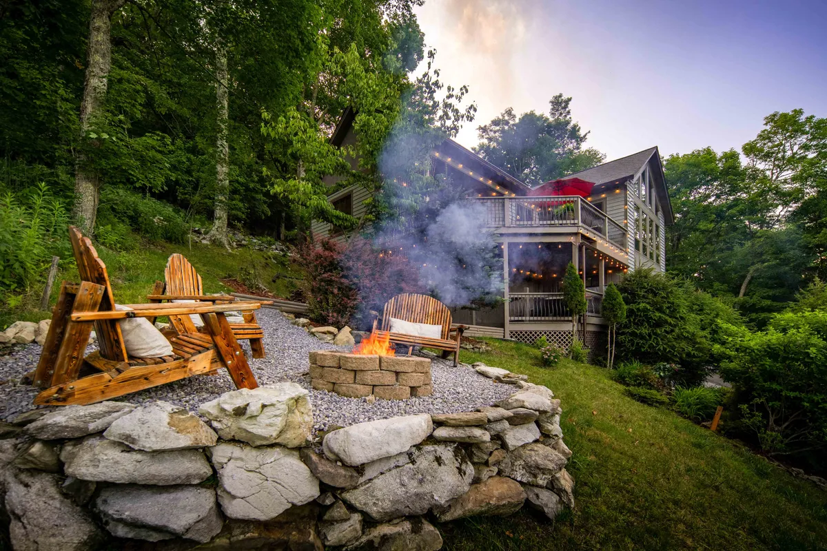 This exterior scene shows a cozy fire pit area with wooden chairs arranged for conversation, all set against the backdrop of the house and lush greenery. The mood is warm and inviting, perfect for storytelling or gathering after dark.
