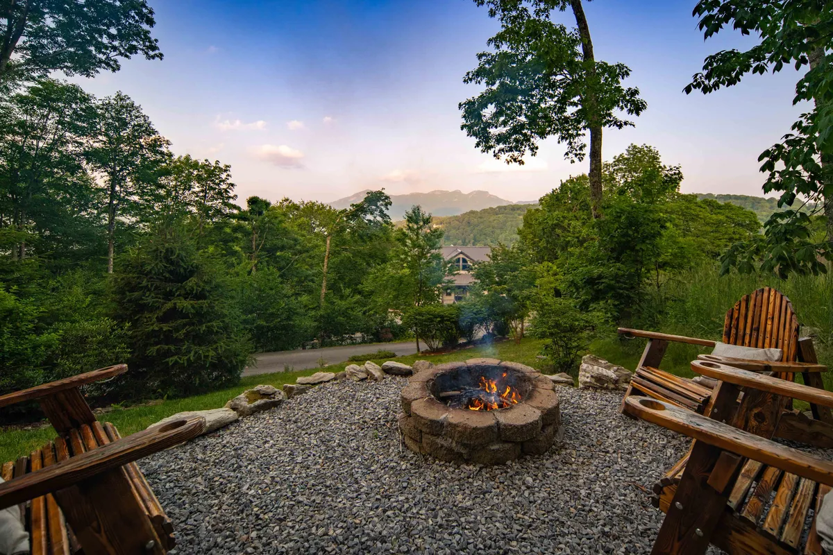 This exterior image showcases a cozy fire pit surrounded by wooden chairs, set against a stunning mountain view. The setting is perfect for groups to gather, share stories, and enjoy the outdoors, especially under the stars or at sunset.