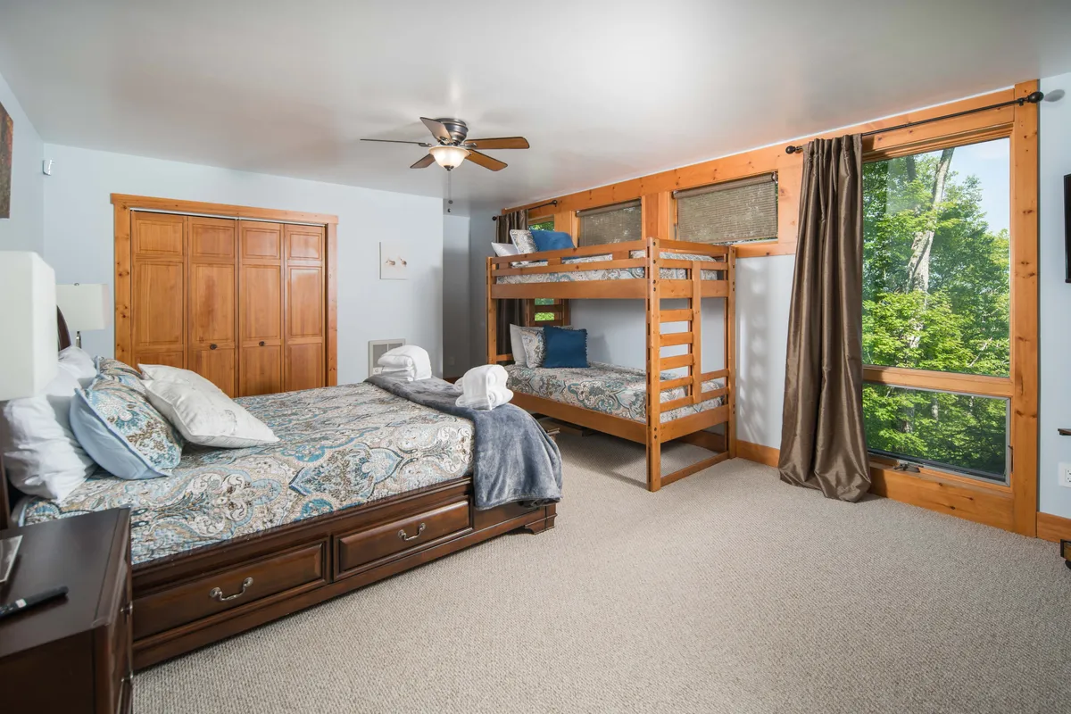 A spacious and inviting bedroom featuring a king-sized bed and a bunk bed, with a large window allowing natural light to fill the space. The wooden furniture and soft linens create a comfortable, family-friendly environment—ideal for accommodating both adults and kids. The overall mood is peaceful and welcoming.