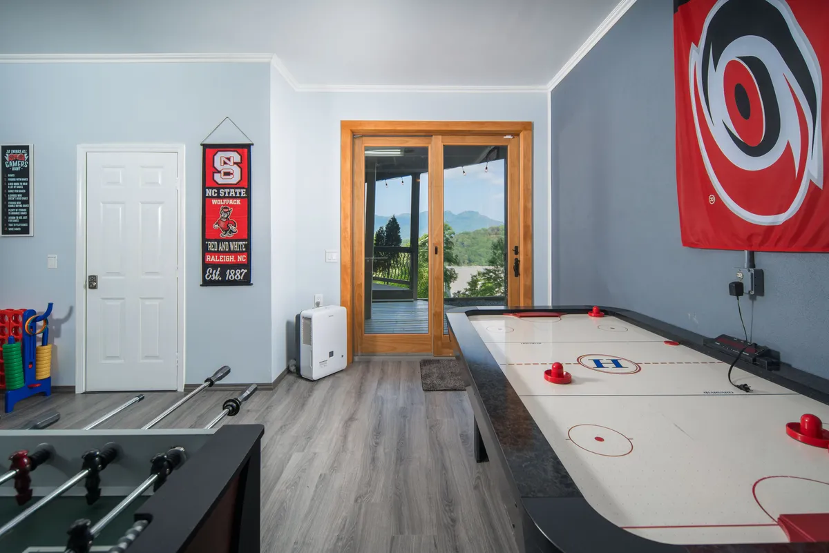 A lively game room displaying both an air hockey table and a foosball table, with double doors leading directly to the patio and an NC State flag adding a fun touch. Sunlight streams in, connecting the indoor activity hub seamlessly to the outside.