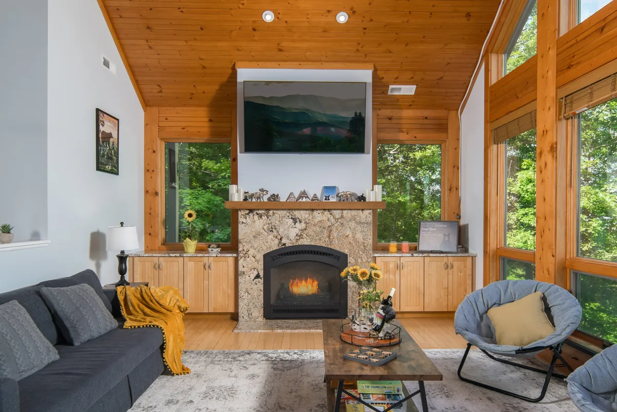A warm and inviting living room featuring a gas log fireplace, a large wall of windows that let in abundant natural light, cozy seating including a sofa, and a coffee table. The windows reveal beautiful mountain vistas, and the arrangement encourages socializing and relaxation.