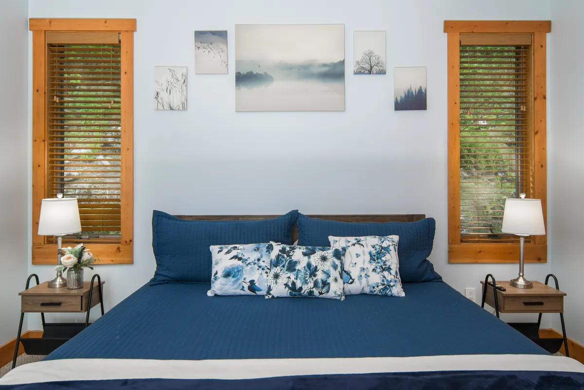 A serene king bedroom with plush blue bedding, decorative pillows, wall art, and nightstands topped with lamps. Sunlight gently filters through, and the tranquil mood invites guests to unwind in privacy and luxury.