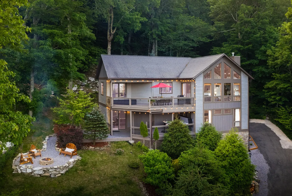 The image showcases the inviting exterior of a single-family home nestled in the trees, featuring a spacious balcony with a striking red umbrella, a cozy fire pit area perfect for gatherings, and the natural beauty of Sugar Mountain in the background. The scene is bathed in the soft glow of sunset, suggesting opportunities for outdoor relaxation, stargazing, and enjoying the serene mountain air.