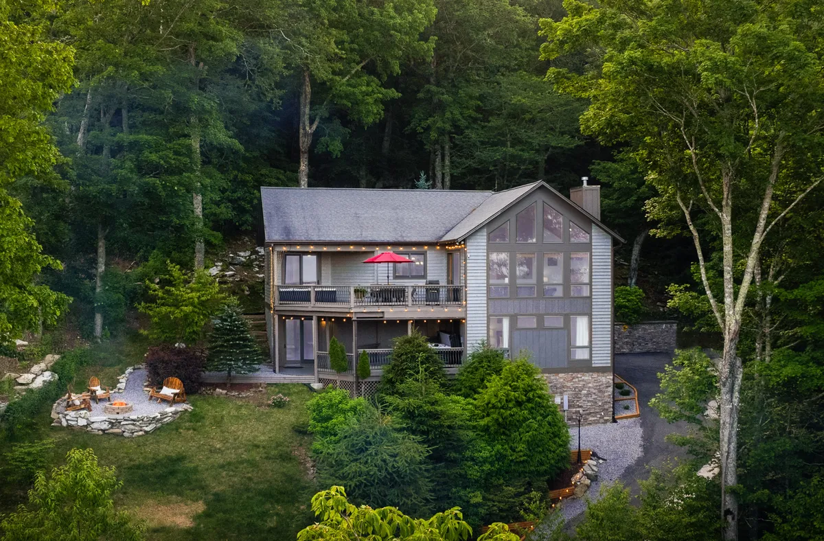 An inviting exterior shot featuring the house facade, a fire pit area, lush greenery, and a patio with a red umbrella. The scene suggests a welcoming atmosphere for outdoor gatherings, barbecues, and shared meals in a natural setting.